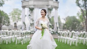 A wedding ceremony with a bride in a white dress holding a bouquet, seated at a table with white chairs, in front of a gazebo.