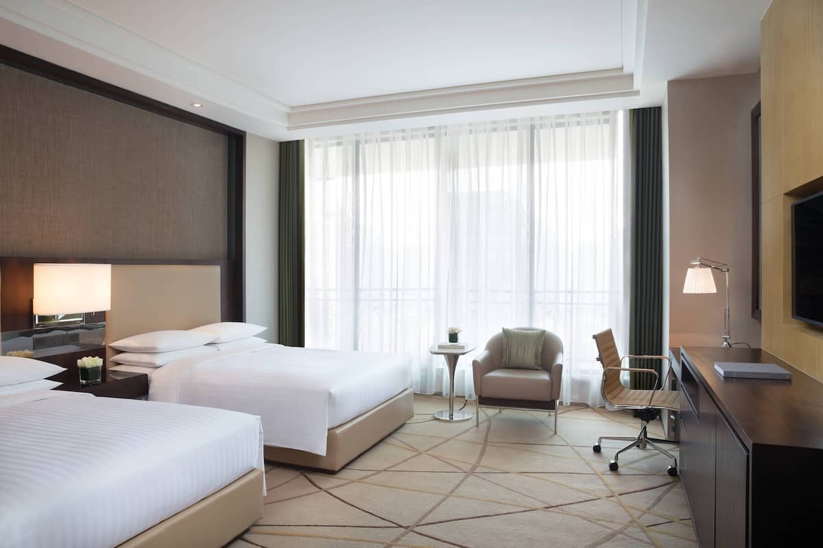 A hotel room with two beds, a desk, a chair, a TV, and a large window with curtains.