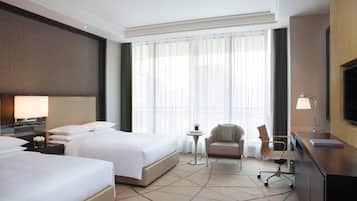 Habitación de hotel con dos camas, un escritorio, una silla, un televisor y un ventanal con cortinas.