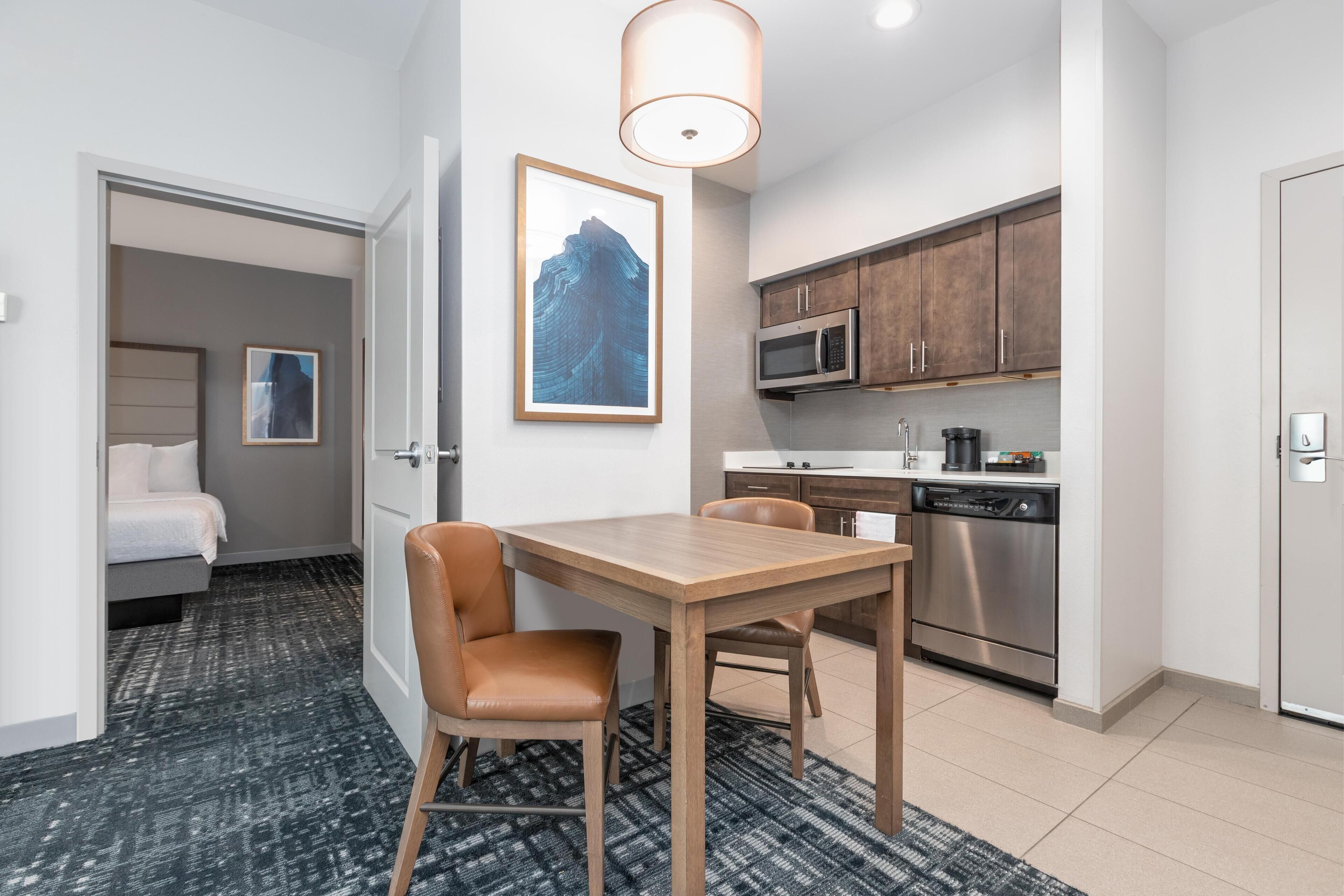 Une chambre d’hôtel moderne avec une table à manger en bois, deux fauteuils en cuir, un coin cuisine équipé d’appareils électroménagers en acier inoxydable et une œuvre d’art encadrée au mur.