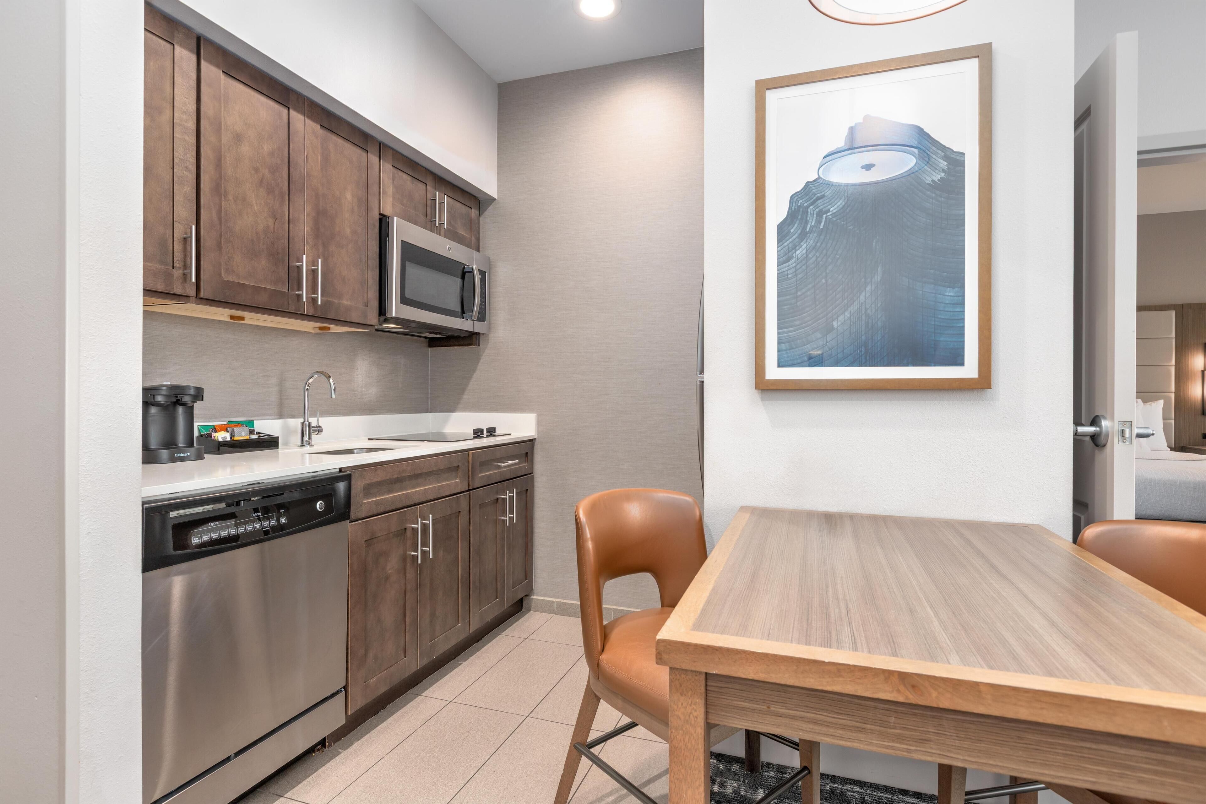 A compact kitchen with stainless steel appliances, a dishwasher, and a microwave. There is a wooden table and two chairs. A framed picture hangs on the wall.