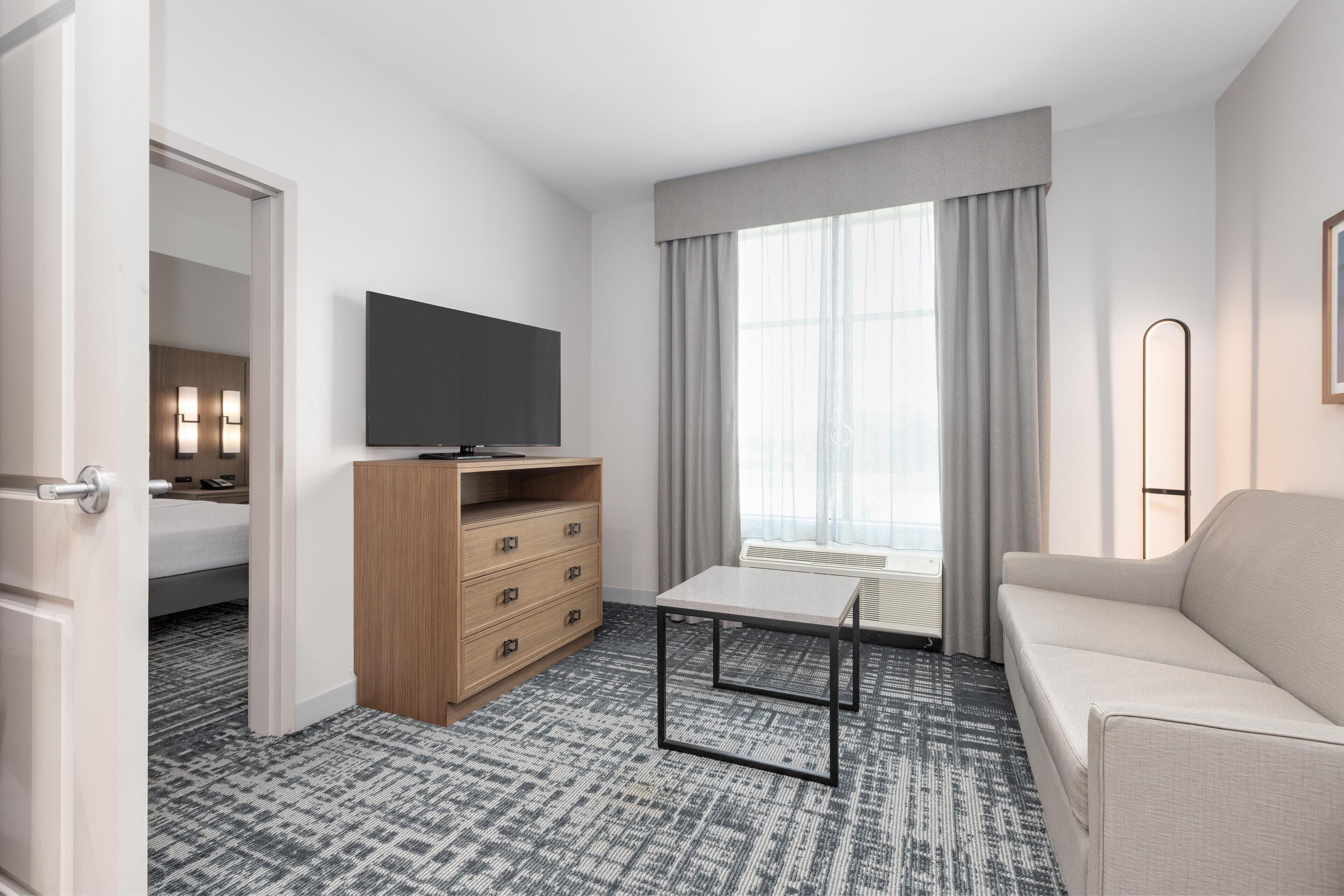 Habitación de hotel con cama, televisión, cómoda de madera, mesa, sofá y ventana con cortinas.