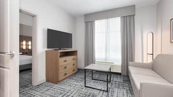 Habitación de hotel con cama, televisión, cómoda de madera, mesa, sofá y ventana con cortinas.