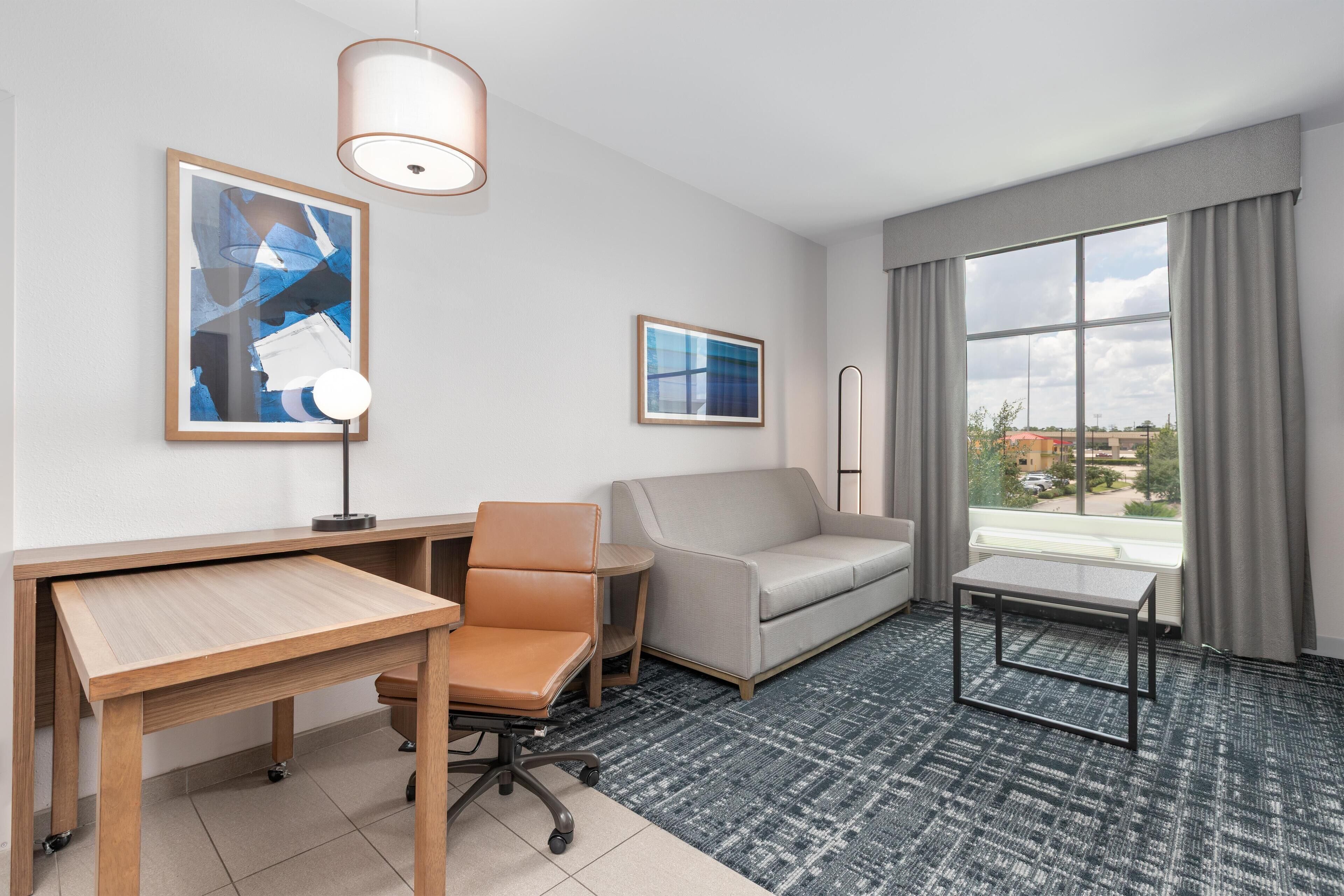 A hotel room with a desk, office chair, sofa, and a small table. There are two framed pictures on the wall, a window with curtains, and a view of buildings outside.