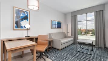 A hotel room with a desk, office chair, sofa, and a small table. There are two framed pictures on the wall, a window with curtains, and a view of buildings outside.