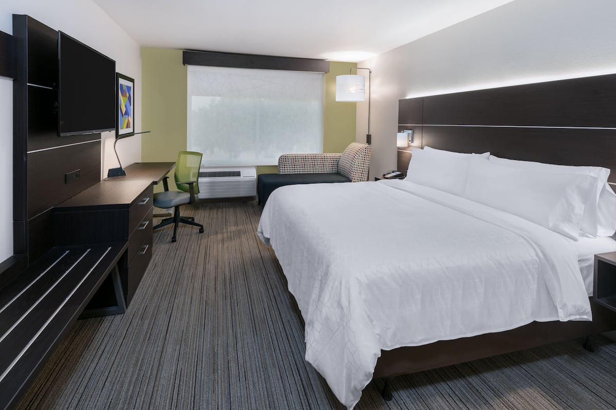 A hotel room with a large bed, a desk with a chair, a flat-screen TV, a window with blinds, and a wall-mounted lamp.