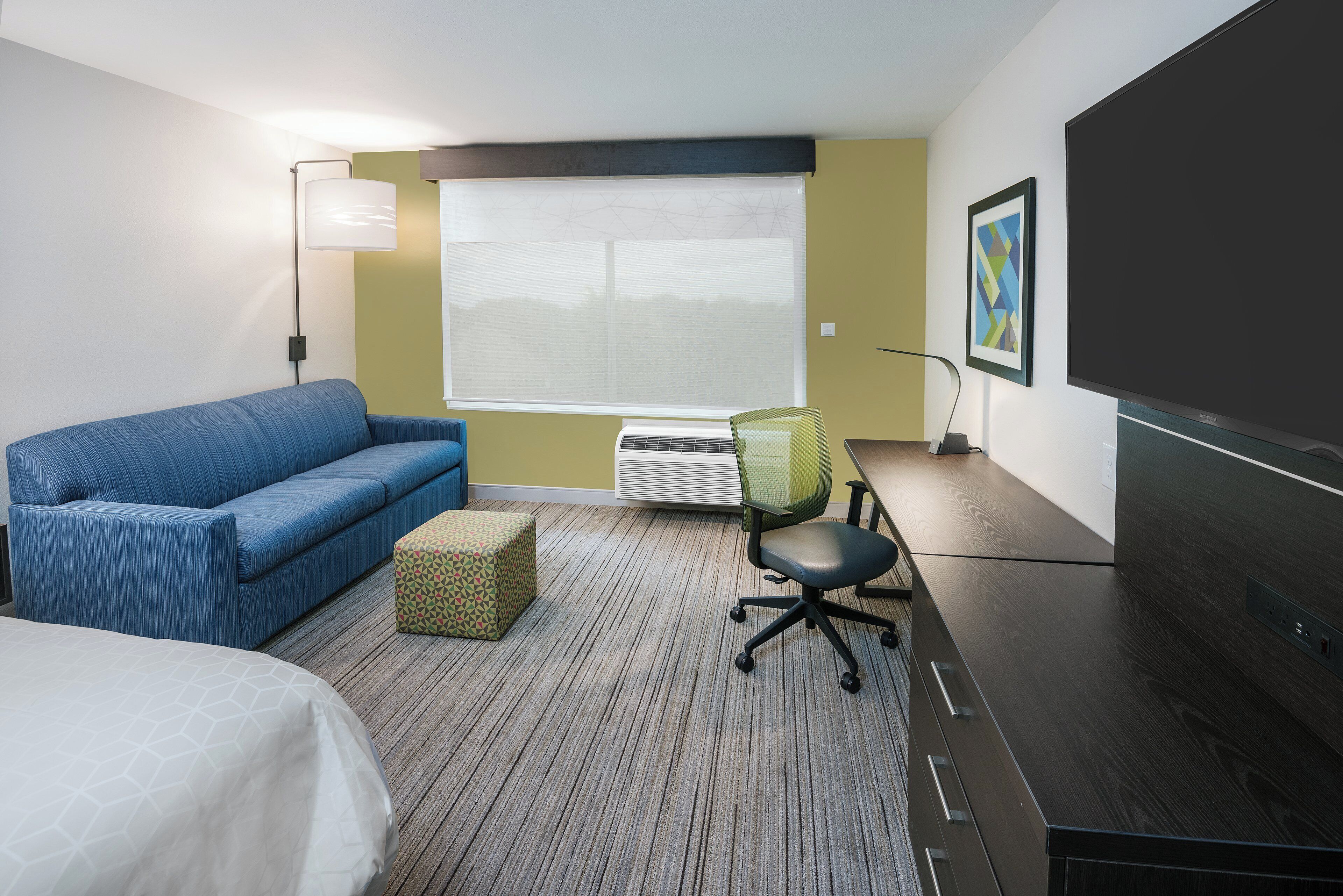 Habitación de hotel con cama, sofá azul, pequeño taburete, escritorio con silla, ventana grande con persianas y televisor de pantalla plana.