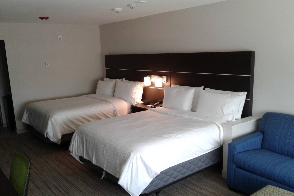 A hotel room with two beds, a nightstand with a lamp, a blue armchair, and a small table.