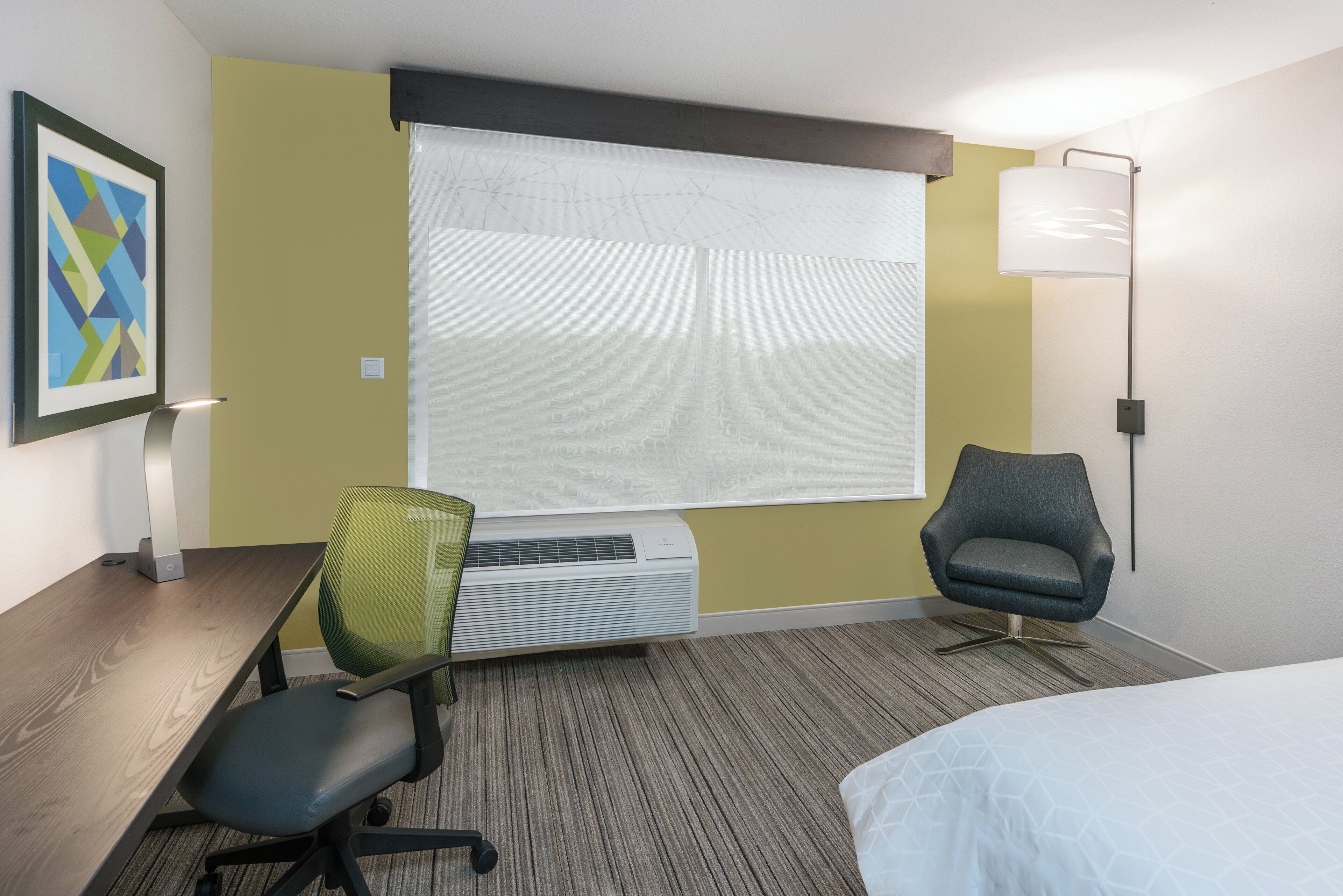 A hotel room with a bed, a desk with a chair, an air conditioning unit, a window with blinds, a lamp, and a framed picture on the wall.