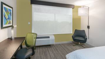 Habitación de hotel con cama, escritorio con silla, lámpara de pie y ventana grande con persianas.