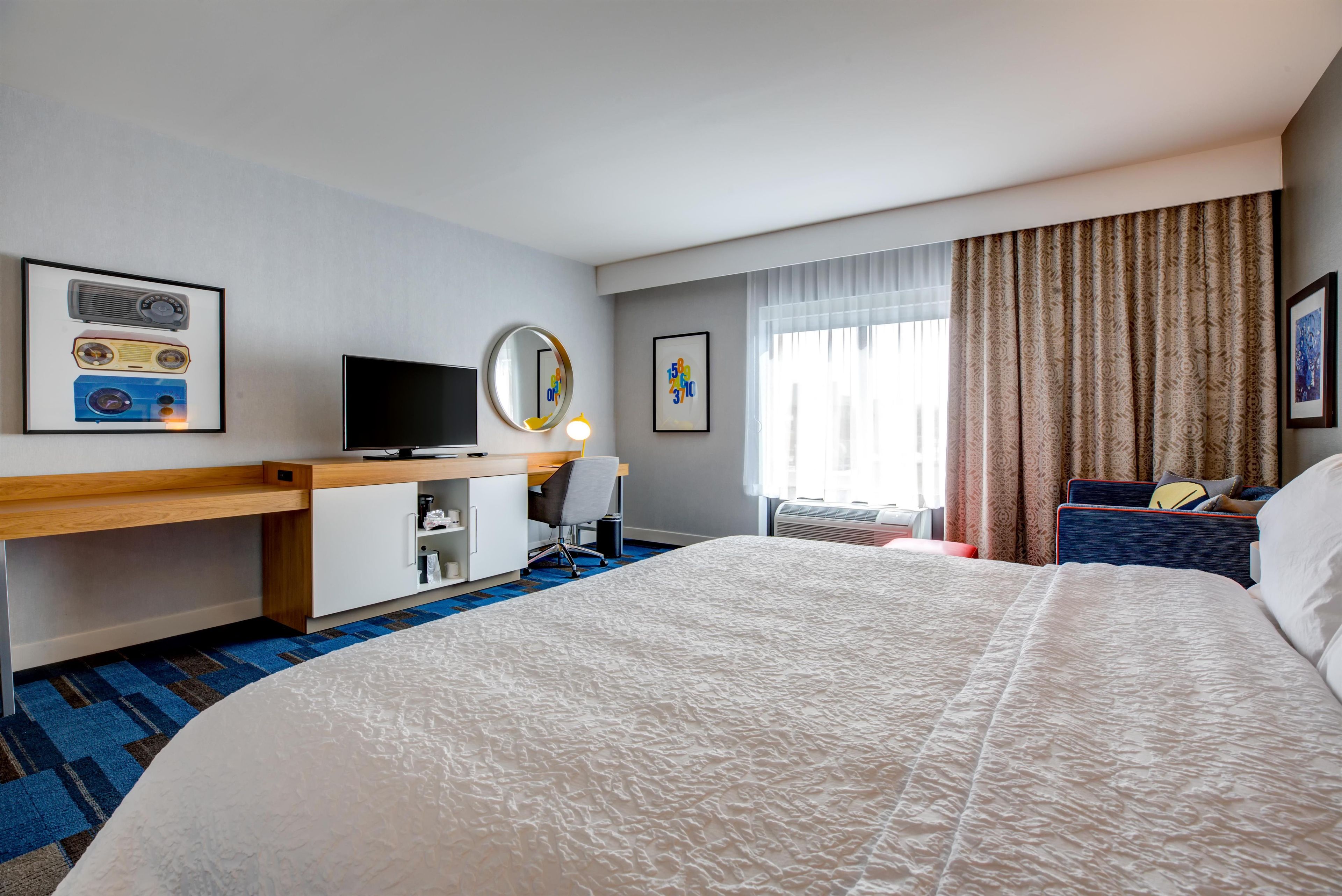 A hotel room with a bed, a desk with a computer, a chair, a mirror, and a window with curtains.
