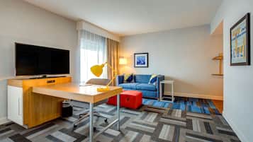 A hotel room with a wooden desk, a flat-screen TV, a blue sofa, a red ottoman, and a patterned carpet.