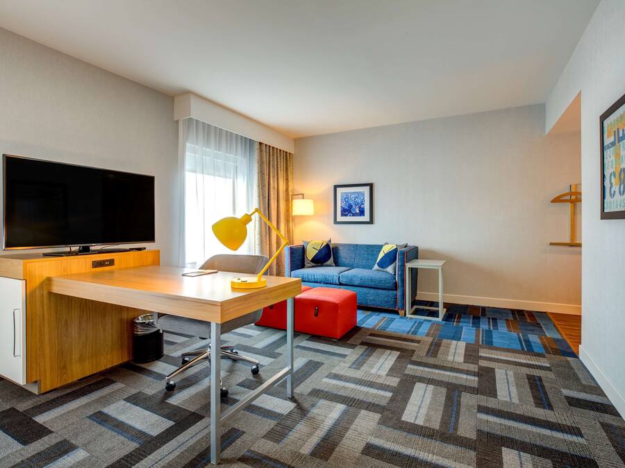 A hotel room with a wooden desk, a flat-screen TV, a blue sofa, a red ottoman, and a patterned carpet.