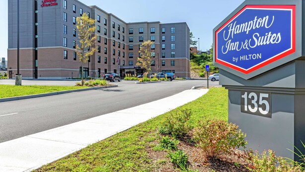 A Hampton Inn & Suites by Hilton building with a clear view of the entrance, parking area, and surrounding greenery.
