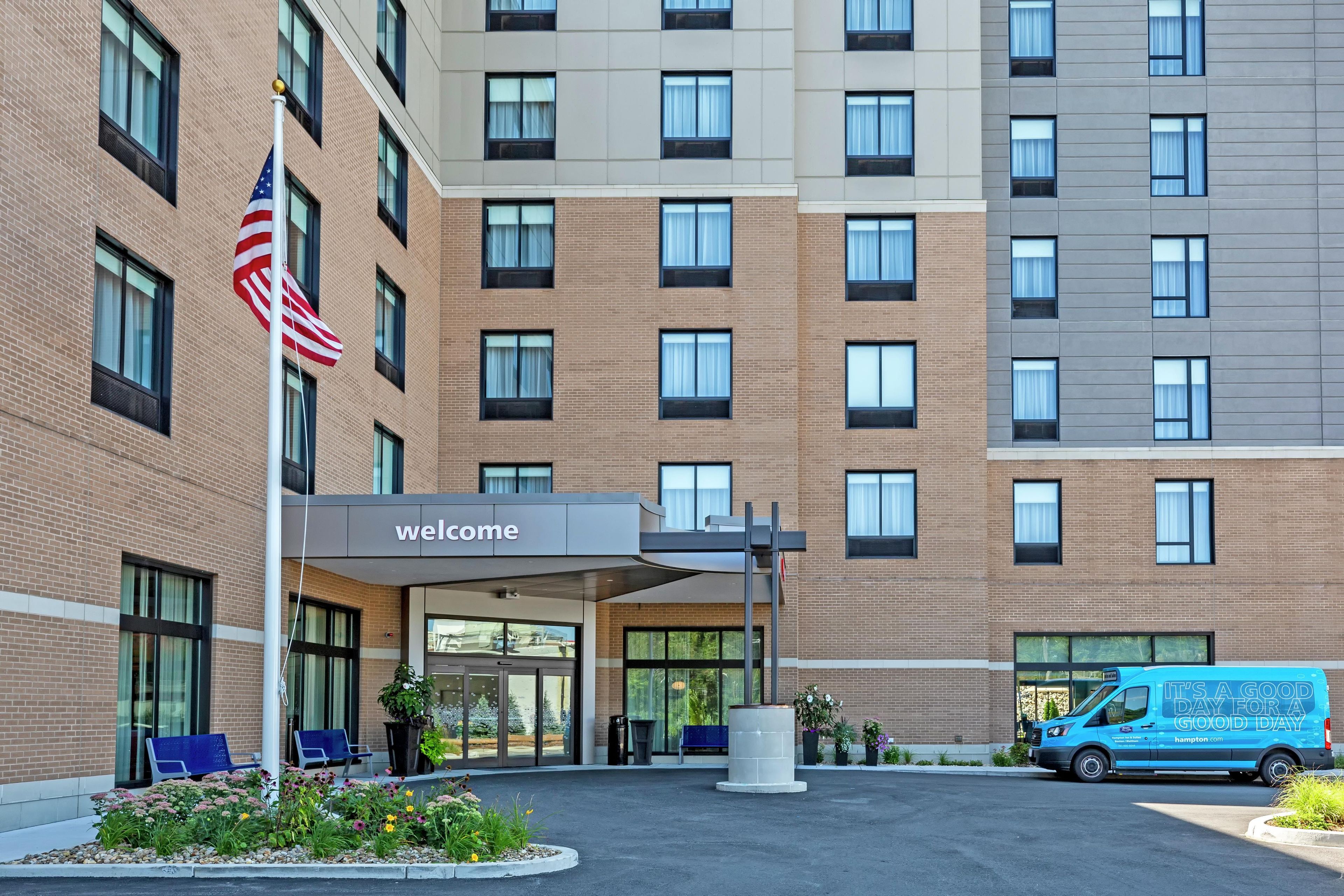 A hotel entrance with a 'welcome' sign, a flag, and a van parked outside.