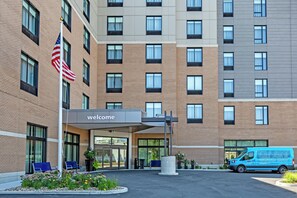 A hotel entrance with a 'welcome' sign, a flag, and a van parked outside. - Hampton Inn & Suites Boston/Waltham (Waltham)