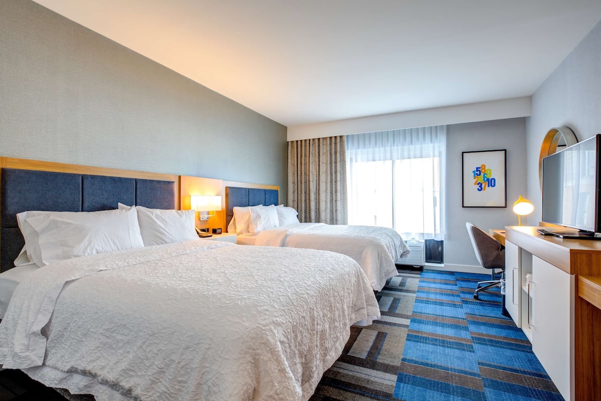 A hotel room with two beds, a desk, a chair, a TV, and a window with curtains.