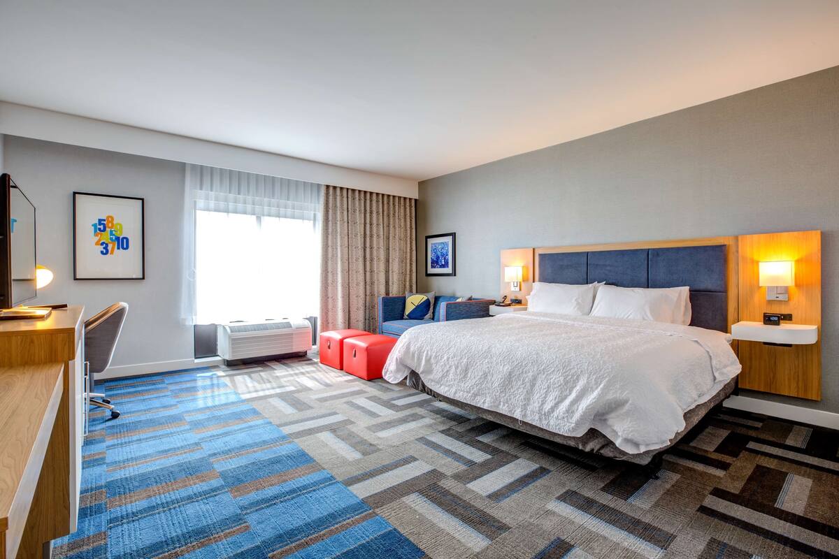A hotel room with a large bed, a desk, a chair, a red ottoman, and a window with curtains.