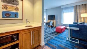 A hotel room with a kitchenette, a flat-screen TV, a sofa, a red ottoman, and a patterned carpet.