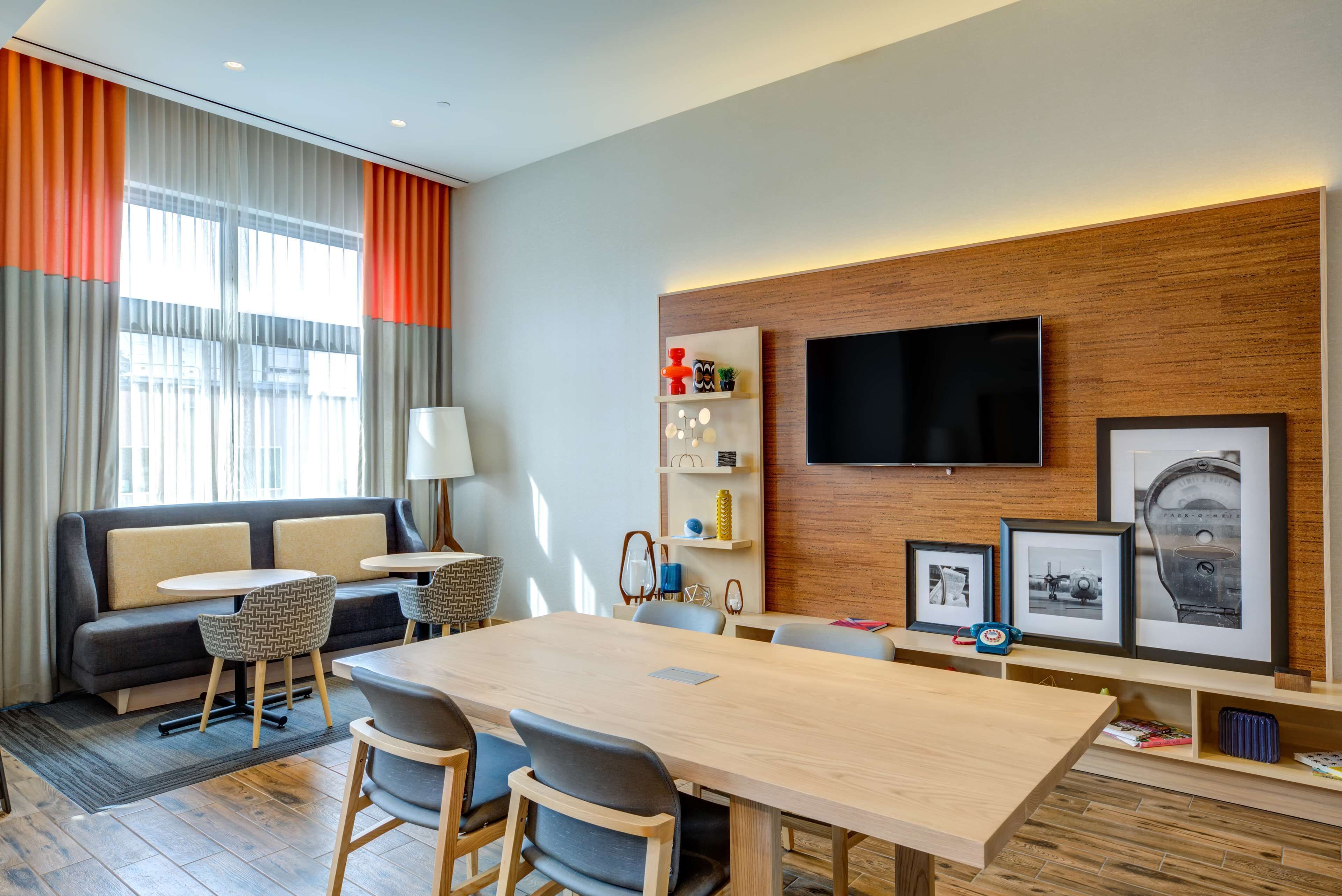 A modern hotel room with a sofa, a dining table, chairs, a TV, and a wooden wall with shelves.
