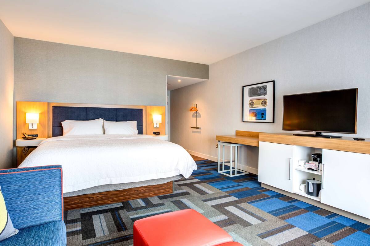 A hotel room with a large bed, bedside lamps, a flat-screen TV, a desk with a chair, and a patterned carpet.