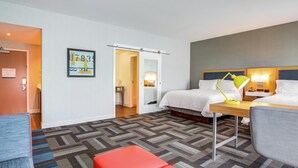 A hotel room with a bed, a desk, a chair, a red ottoman, and a patterned carpet.