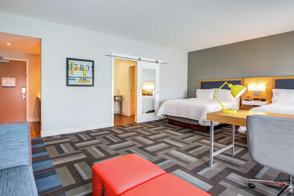 A hotel room with a bed, a desk, a chair, a red bench, a patterned carpet, and a door leading to a bathroom.