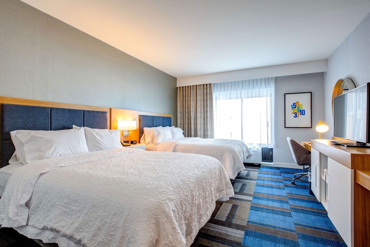 A hotel room with two beds, a desk with a chair, a television, and a window with curtains.