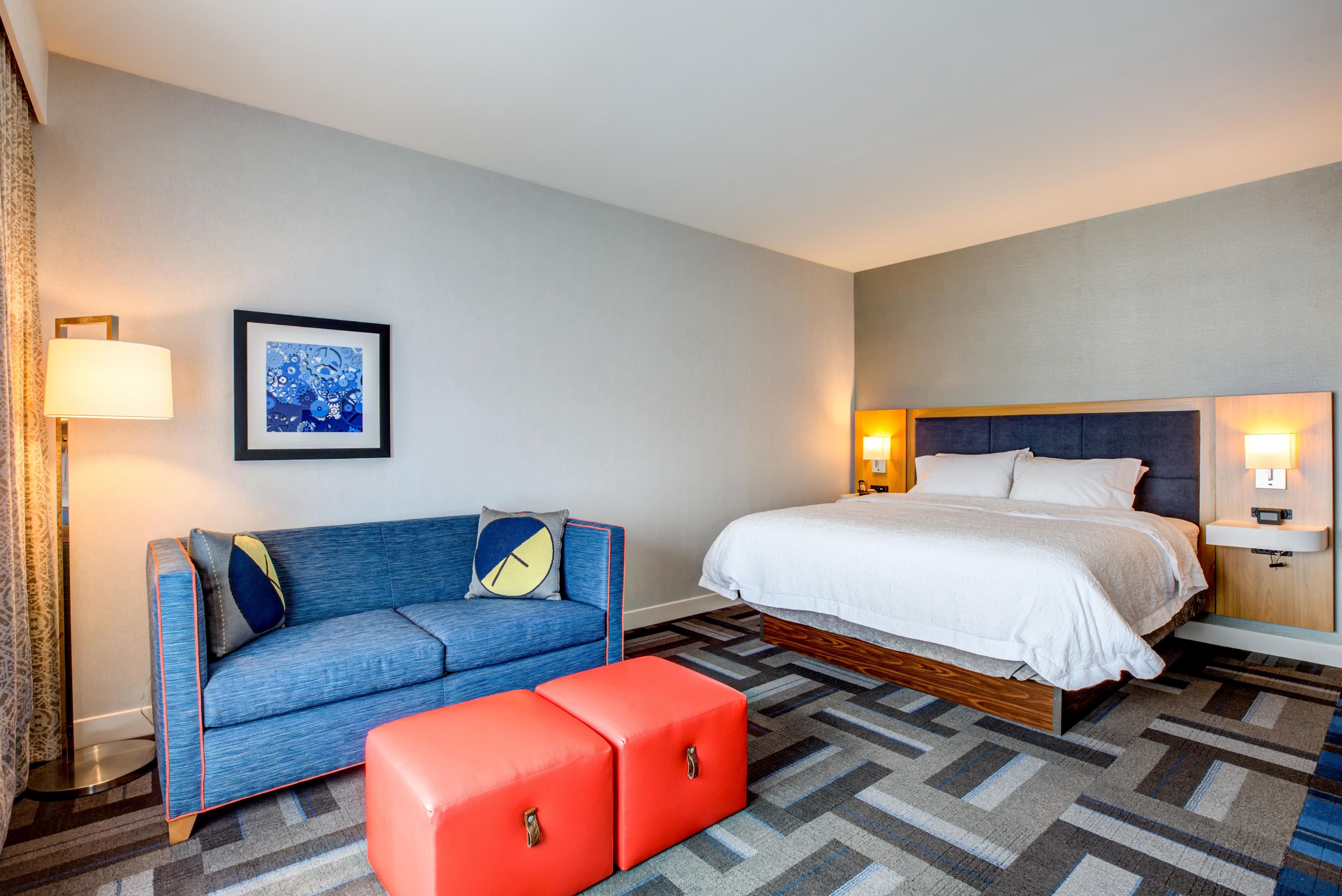 A hotel room with a bed, a sofa, an ottoman, a framed picture on the wall, and two bedside lamps.