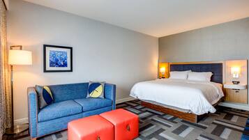 A hotel room with a bed, a sofa, an ottoman, a framed picture on the wall, and two bedside lamps.