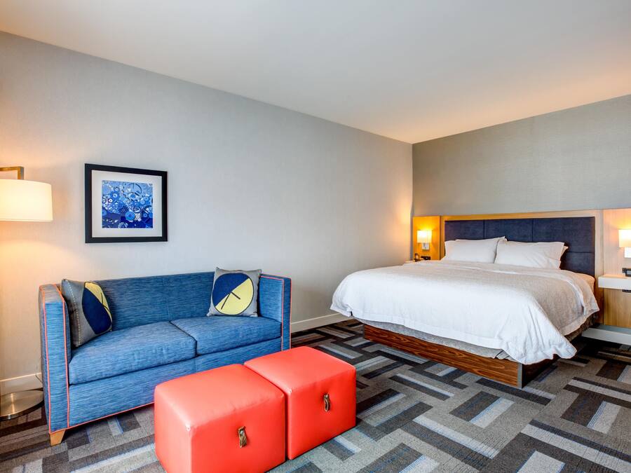Une chambre d’hôtel avec un lit, un canapé, un pouf, un tableau encadré au mur et deux lampes de chevet.
