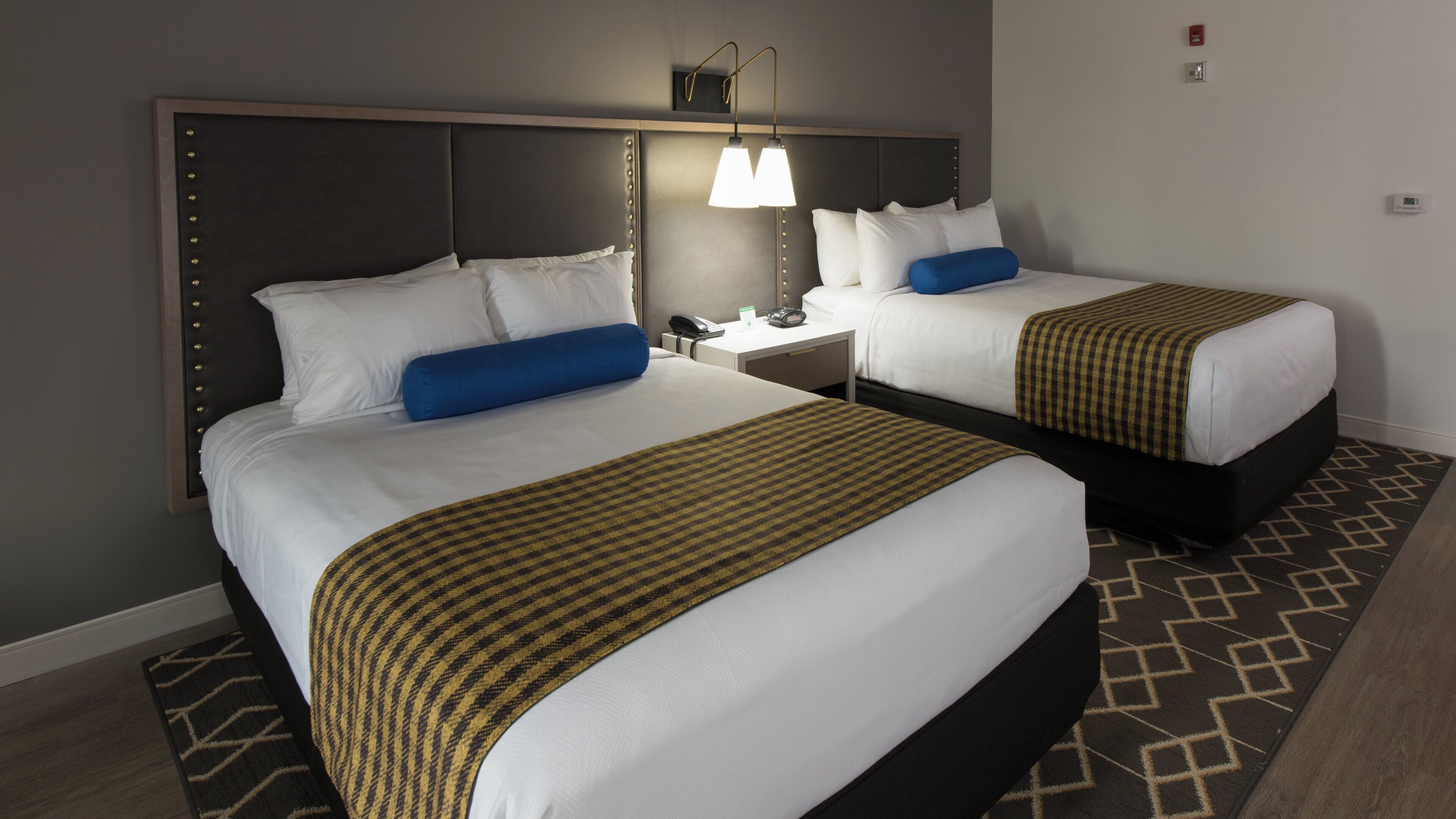 A hotel room with two beds, a nightstand, a lamp, and a patterned rug.