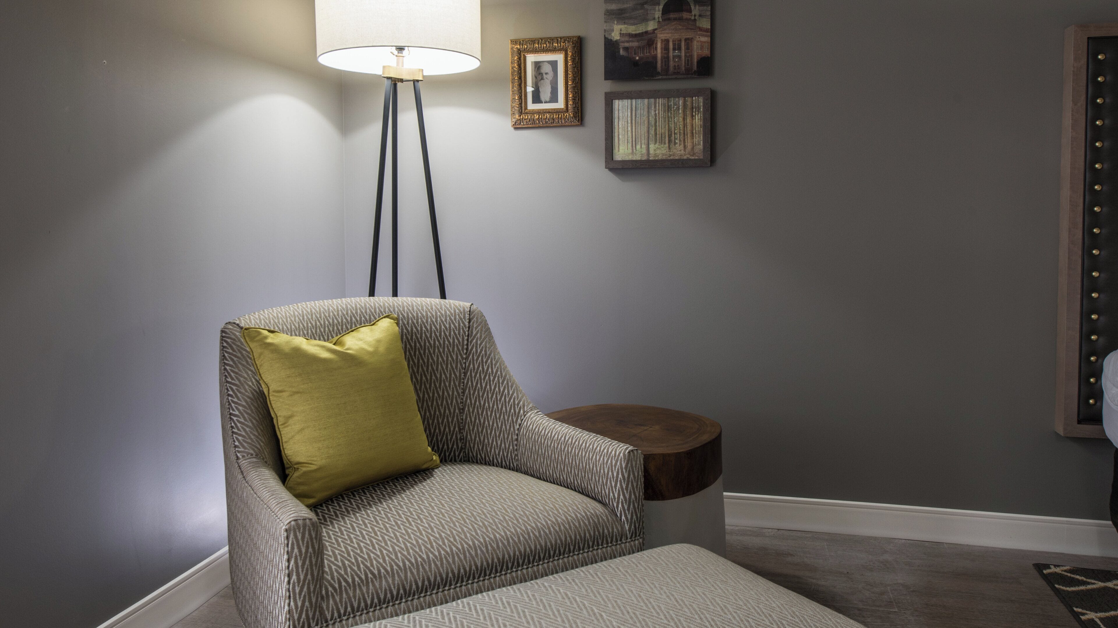 A corner room with a grey armchair, a small round wooden side table, a floor lamp, and two framed pictures on the wall.