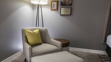 Una habitación con un sillón tapizado en gris, un cojín amarillo a juego, un pequeño taburete, una mesita de madera y una lámpara de pie.