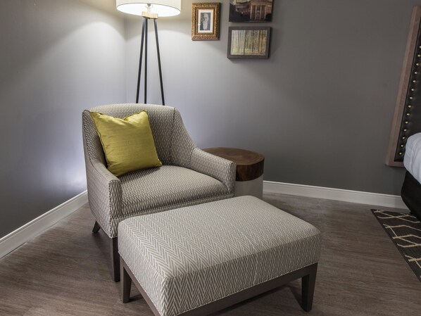 A room with a grey upholstered armchair with a yellow pillow, a matching ottoman, a small wooden side table, and a floor lamp.