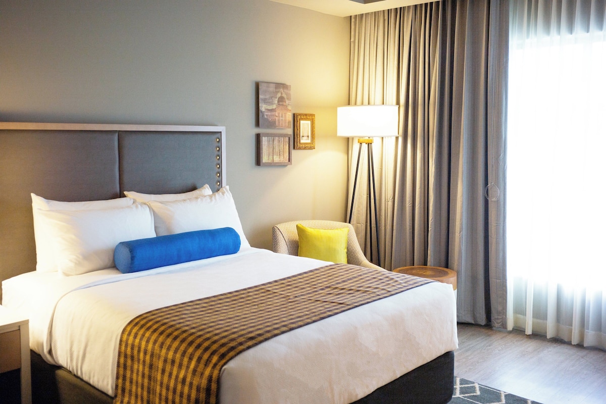 Une chambre d’hôtel avec un grand lit, une table de chevet, une lampe, une chaise et une fenêtre avec des rideaux.