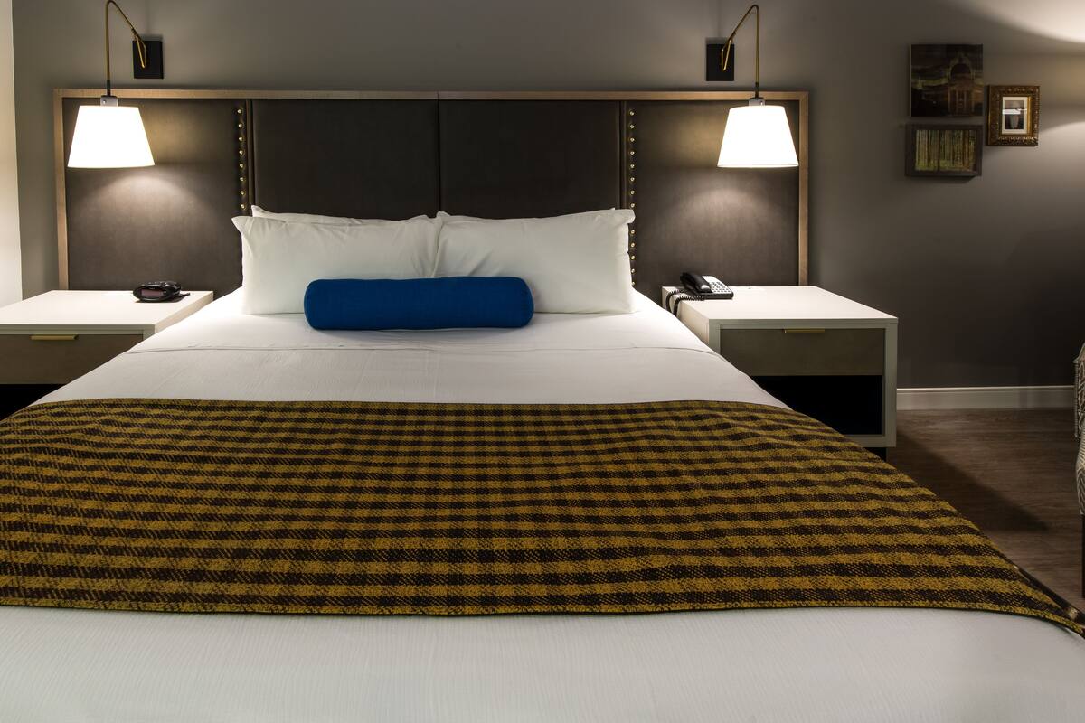 Une chambre d’hôtel avec un grand lit, deux tables de chevet, deux lampes, un oreiller bleu et un plaid à motifs.