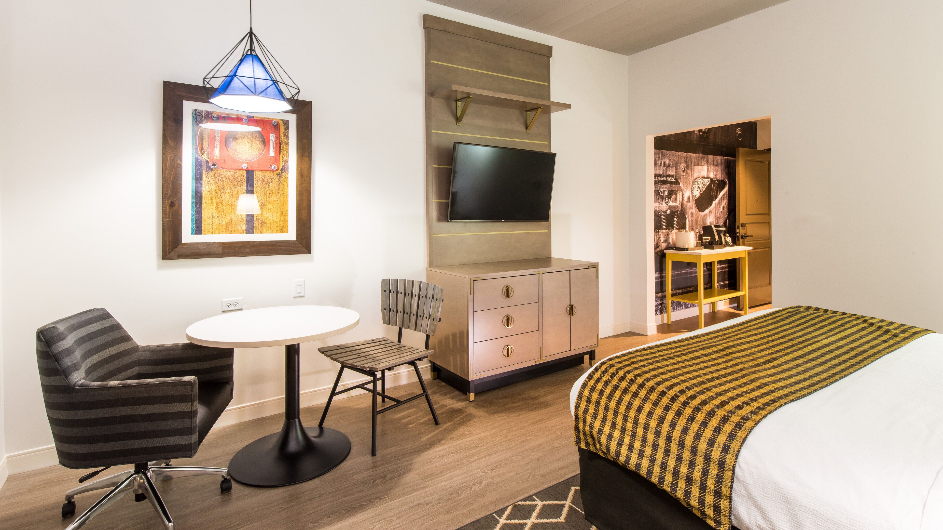 A hotel room with a bed, a desk, a chair, a TV mounted on a wall, and a small table.