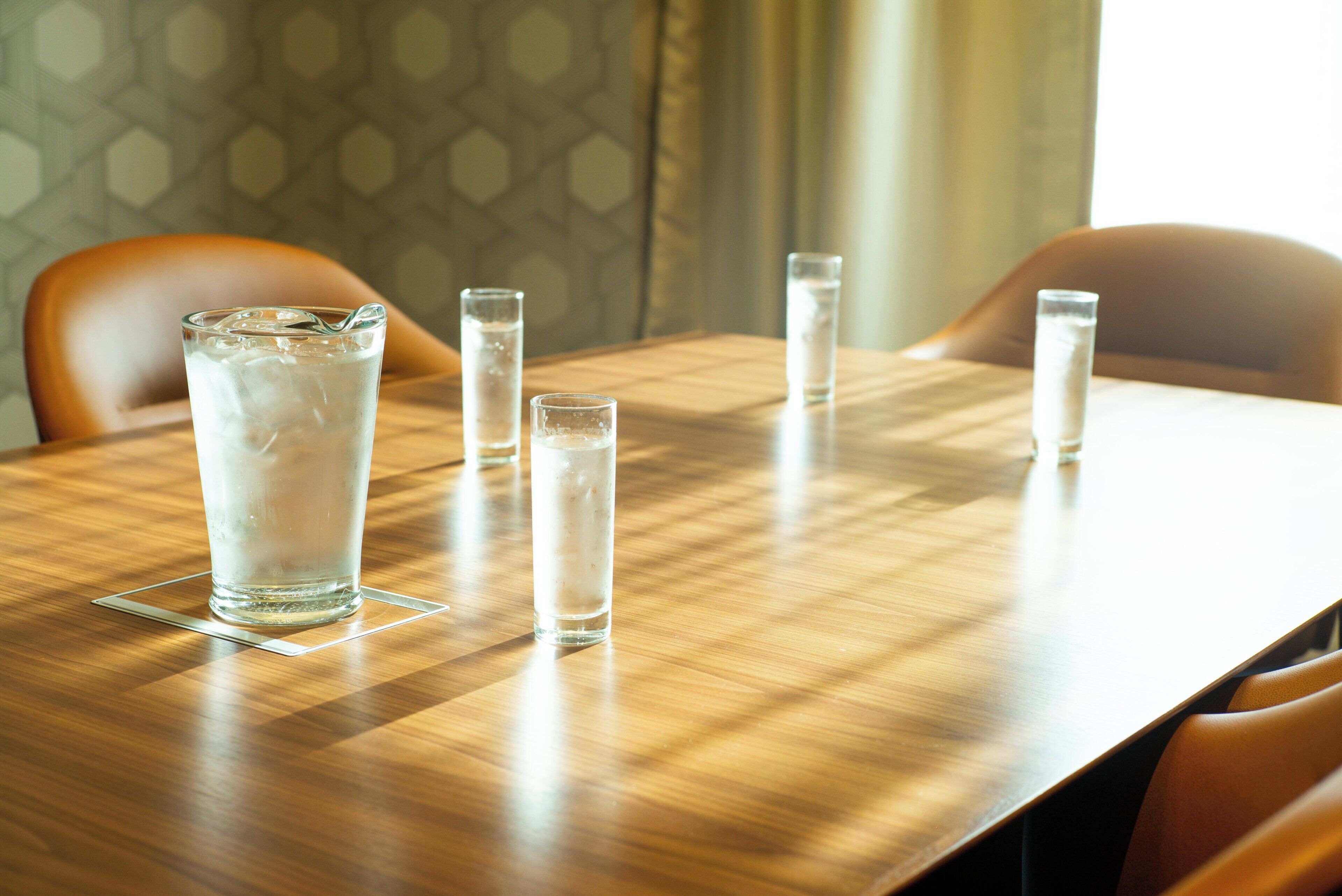 A wooden table with four glasses of water and ice cubes, set in a room with patterned wallpaper and a window with curtains.