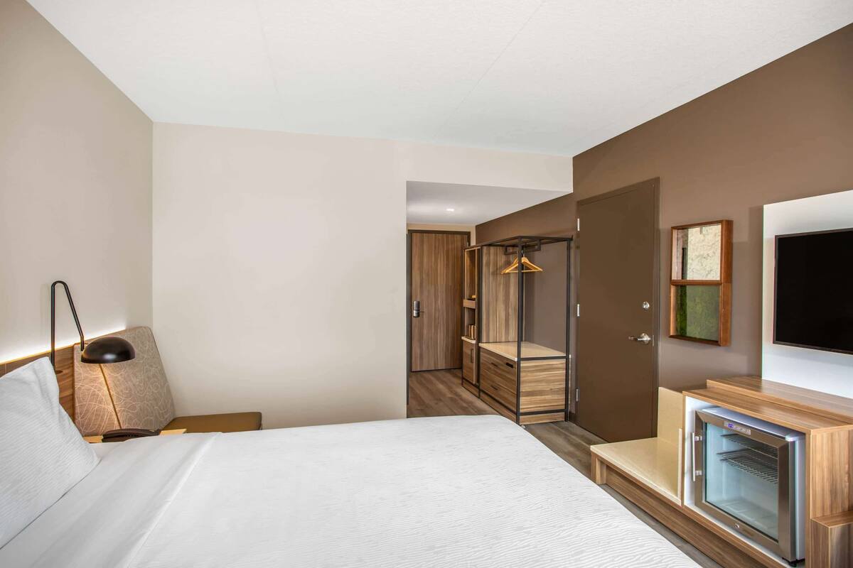Una habitación de hotel con cama, mesita de noche, lámpara, armario, espejo y televisión.