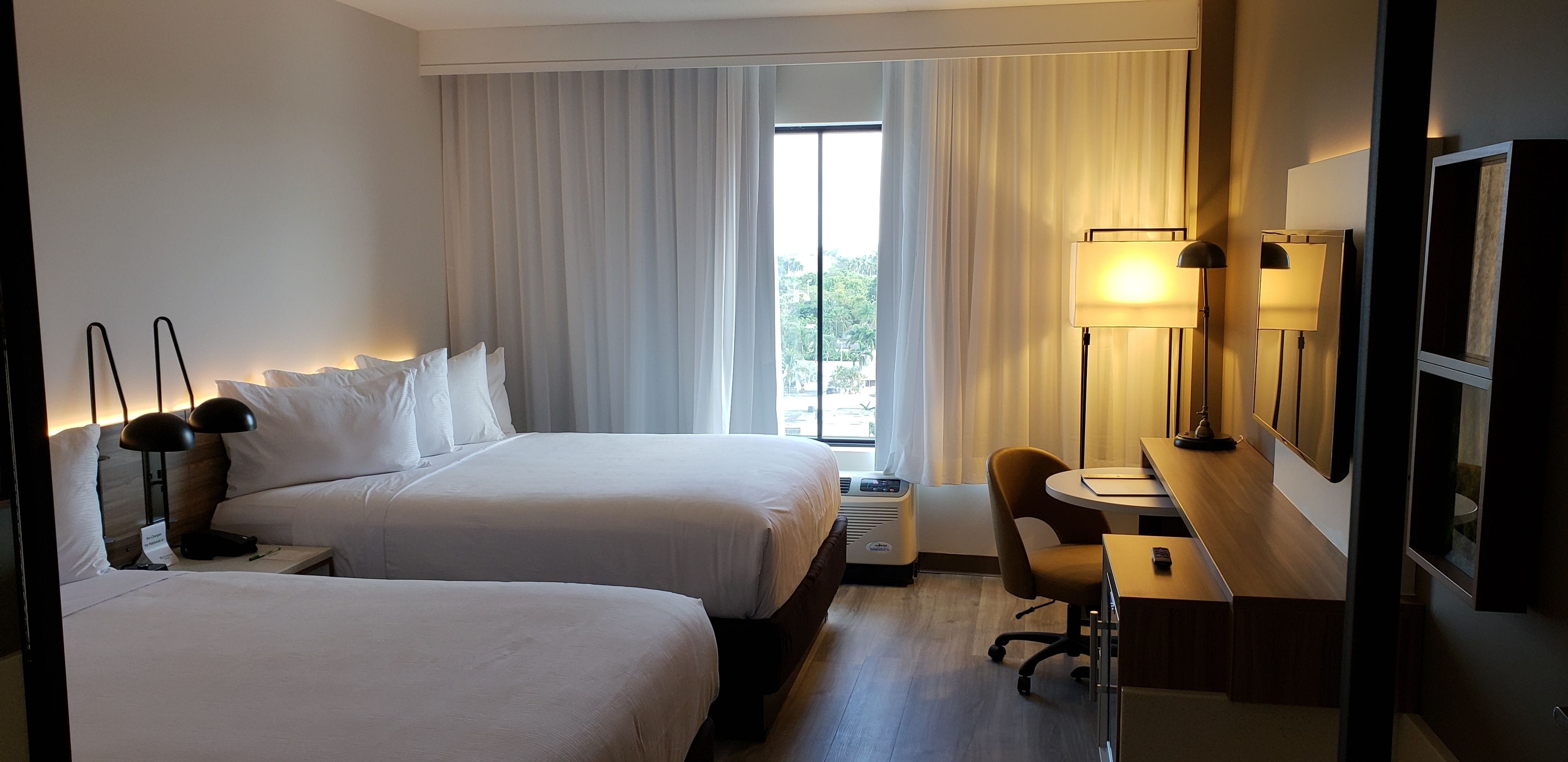 Habitación de hotel con dos camas, un escritorio con silla, lámpara de escritorio, calefactor y una ventana grande con cortinas.