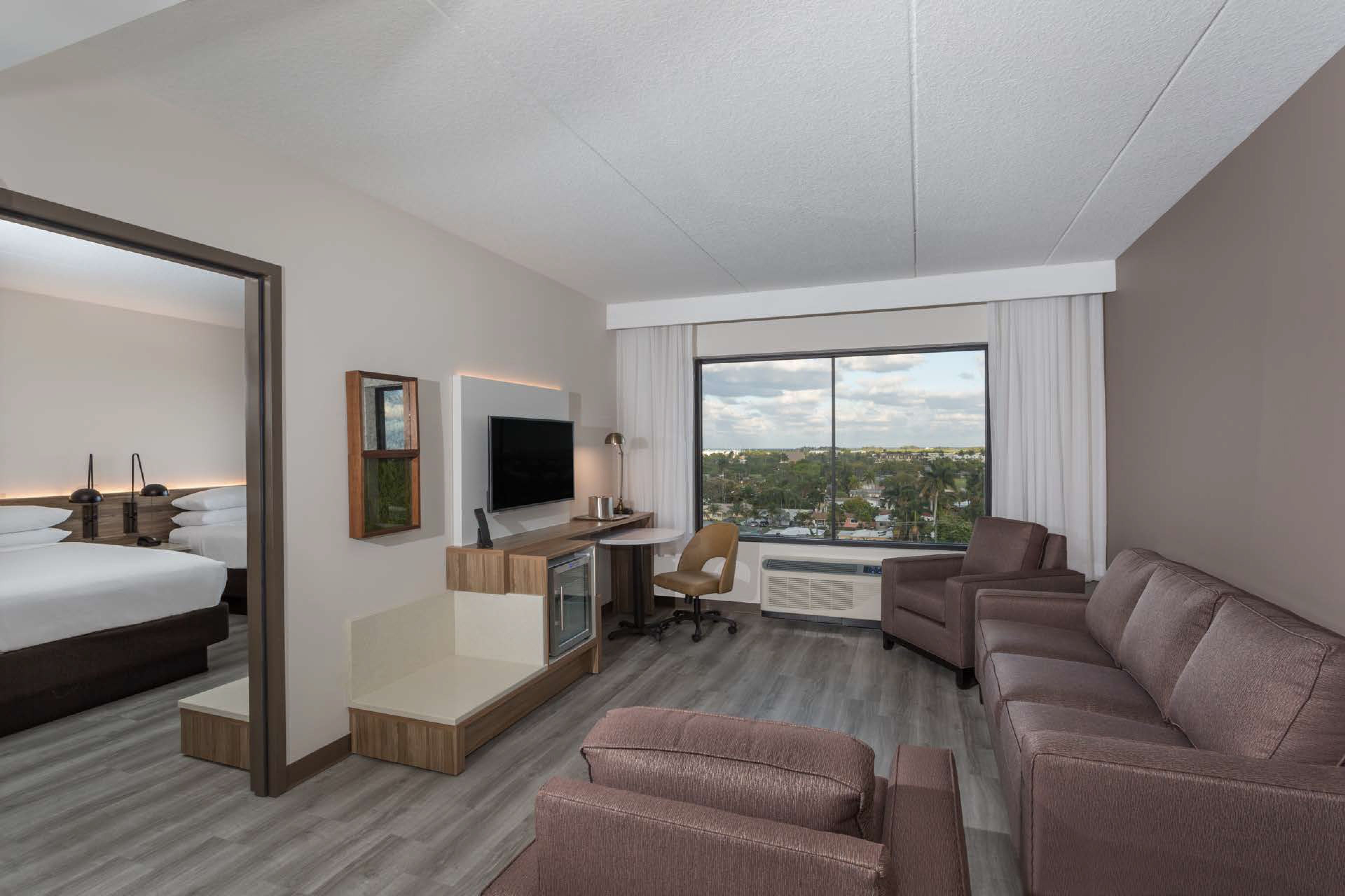 A modern hotel room with a bed, a sofa, a TV, a desk, and a large window offering a city view.