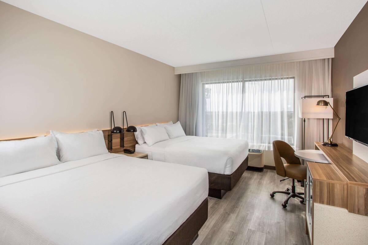 Habitación de hotel con dos camas, un escritorio, una silla, un televisor y un ventanal con cortinas.