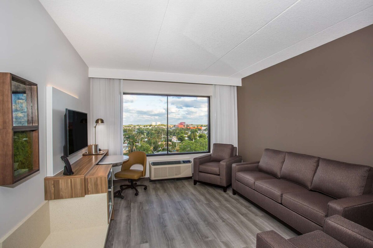 Una sala de estar moderna con un ventanal grande, un televisor de pantalla plana, un sofá, una mesa de centro y una silla.
