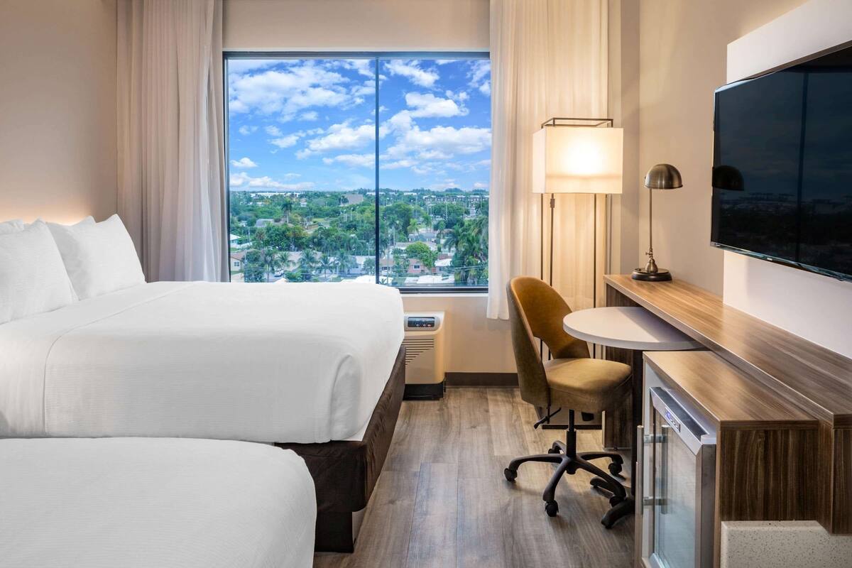 Habitación de hotel con dos camas, un escritorio con lámpara, televisión y una ventana grande con vista a la ciudad.