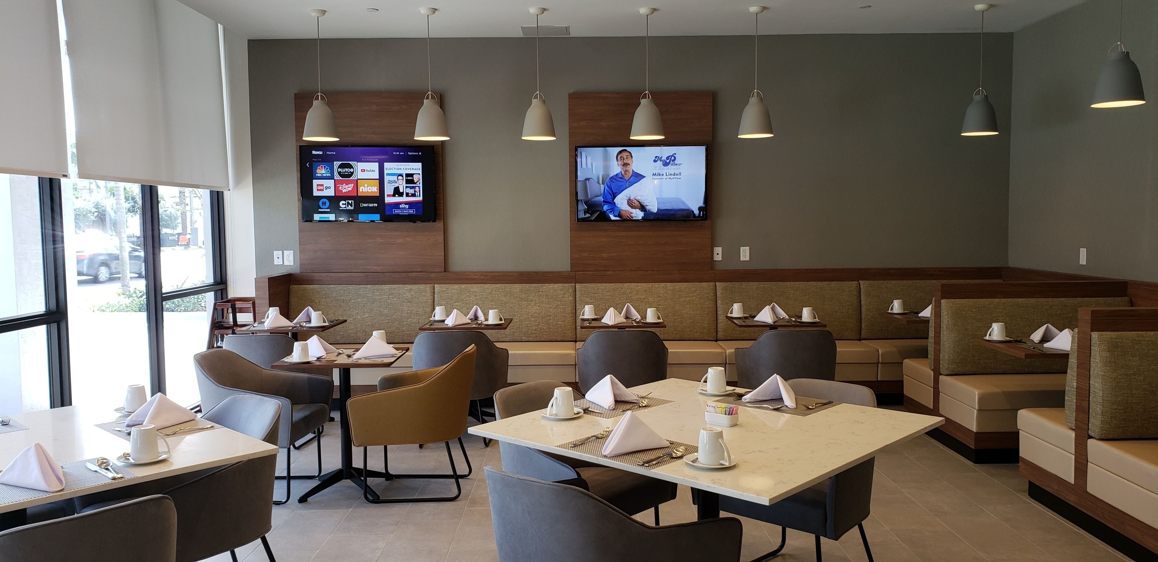 Un restaurante con mesas y sillas, una pantalla de televisión que muestra un noticiero, y vistas al exterior a través de amplios ventanales.
