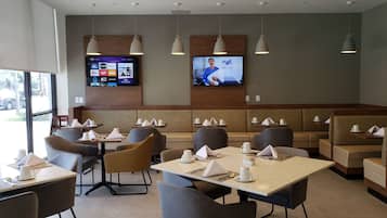 Un restaurante con mesas y sillas, una pantalla de televisión que muestra un noticiero, y vistas al exterior a través de amplios ventanales.