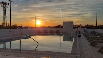 Piscina com cadeiras de descanso e pôr do sol ao fundo.