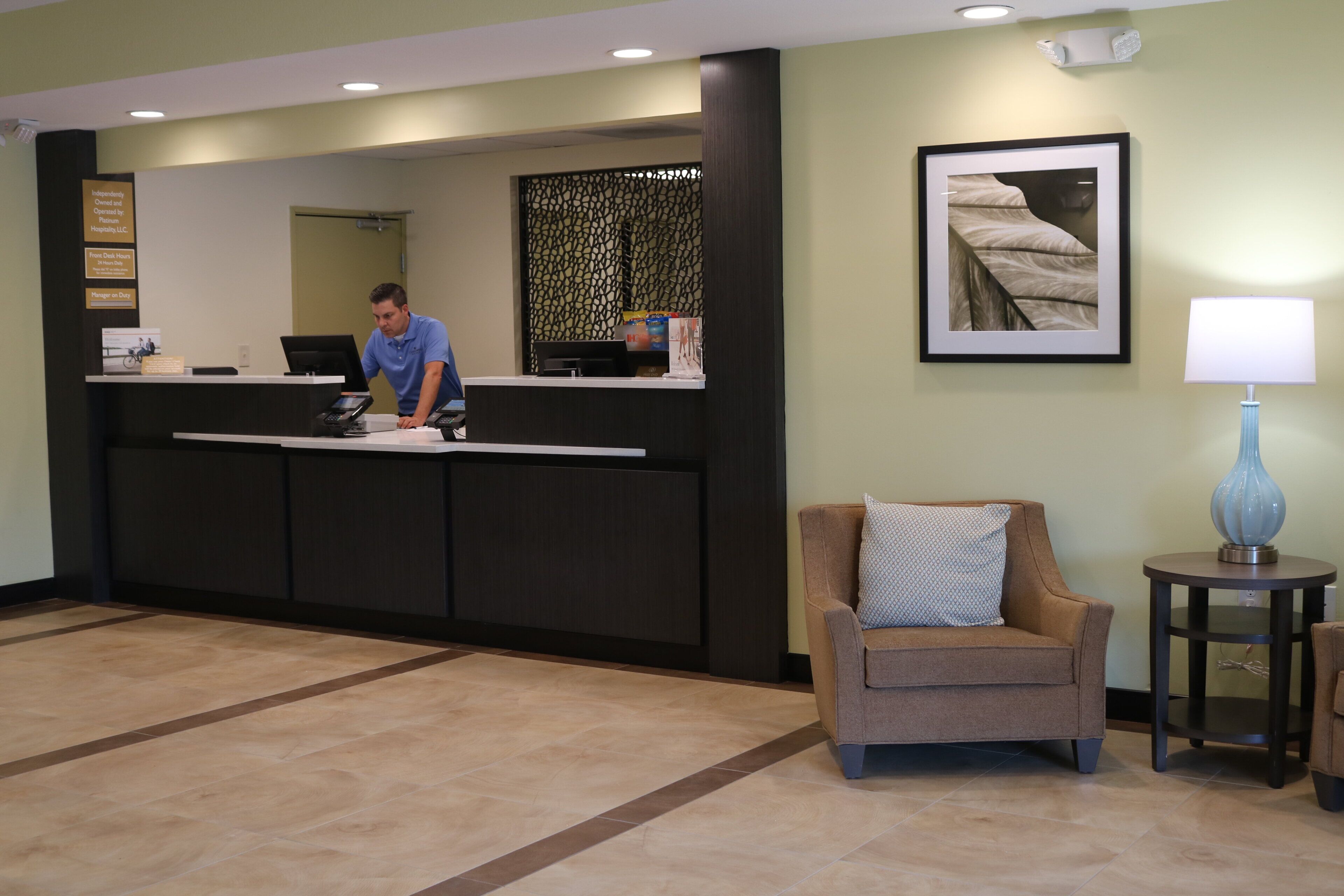 A reception desk with a man standing behind it, a chair, a small table with a lamp, and a framed picture on the wall.