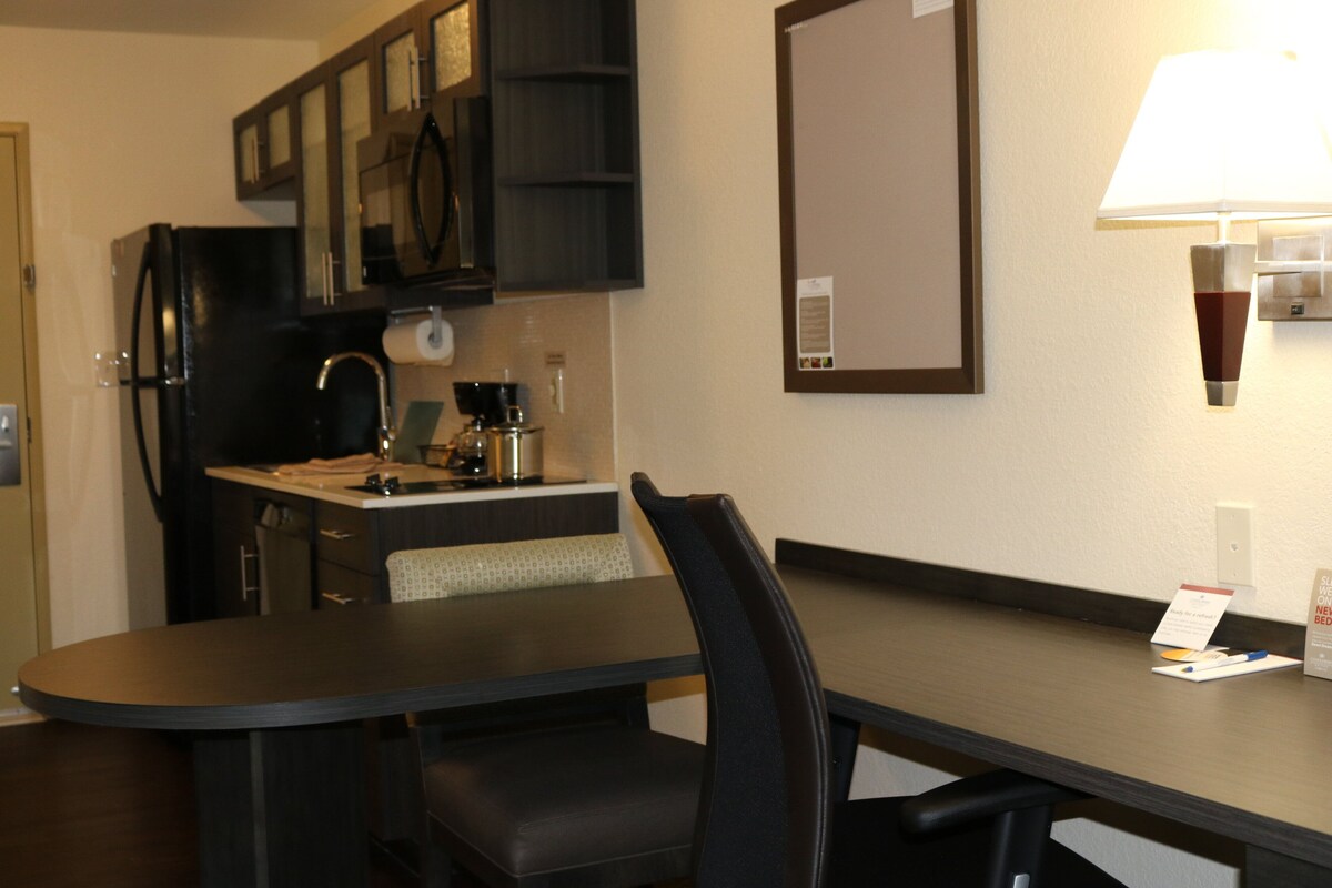 Quarto de hotel com mesa de jantar, cadeiras, geladeira, cafeteira, espelho e abajur.